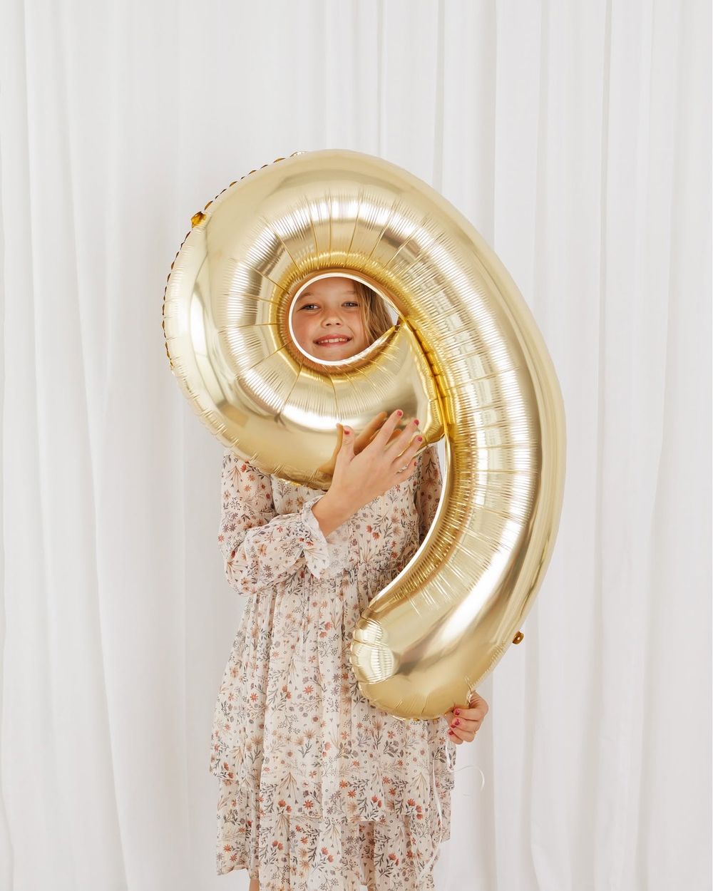 A smiling child in a floral dress holds a large gold number 9 foil balloon in front of a white curtain.