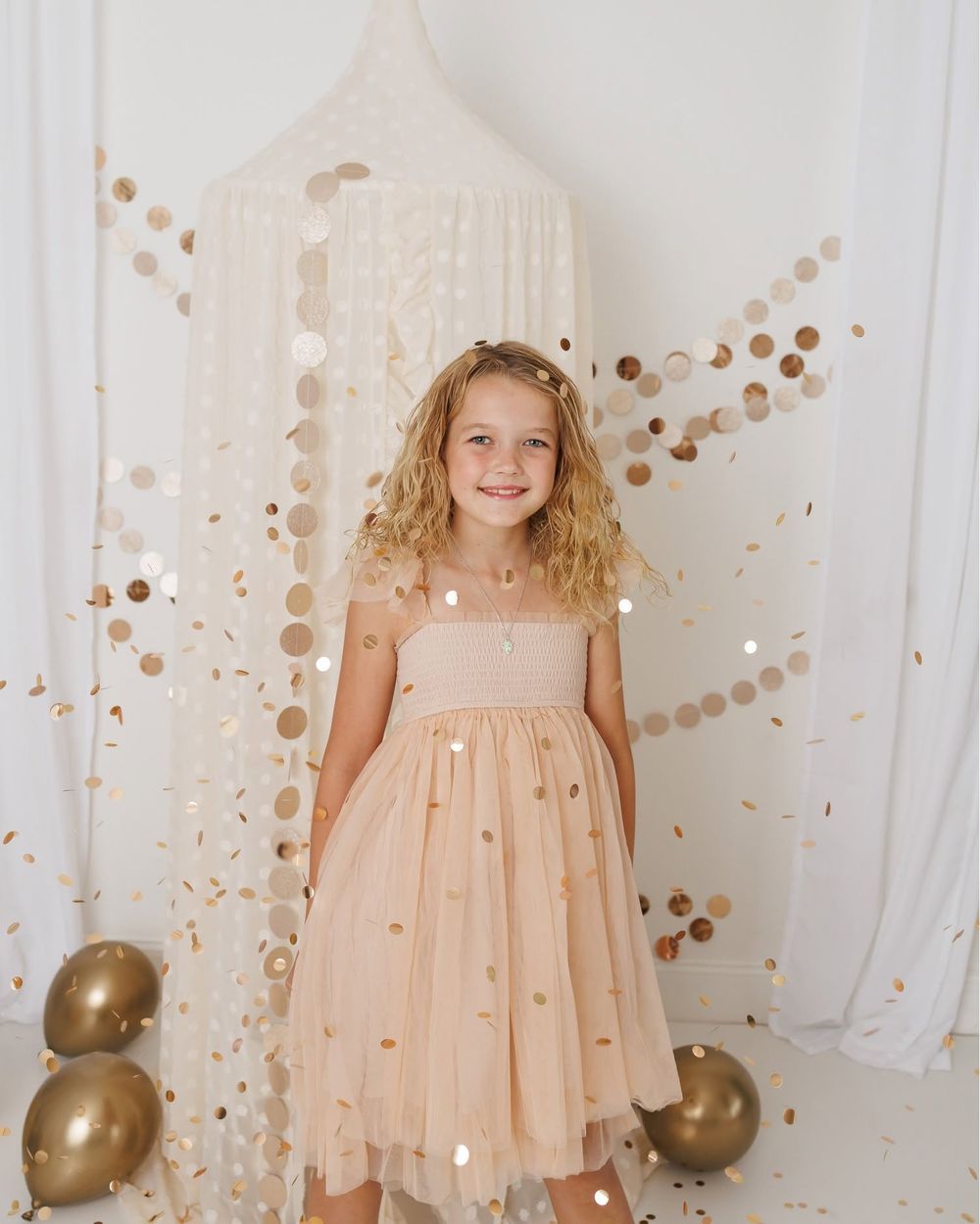 Young girl in peach tulle dress smiles amid gold confetti and balloons in a bright white studio setting.