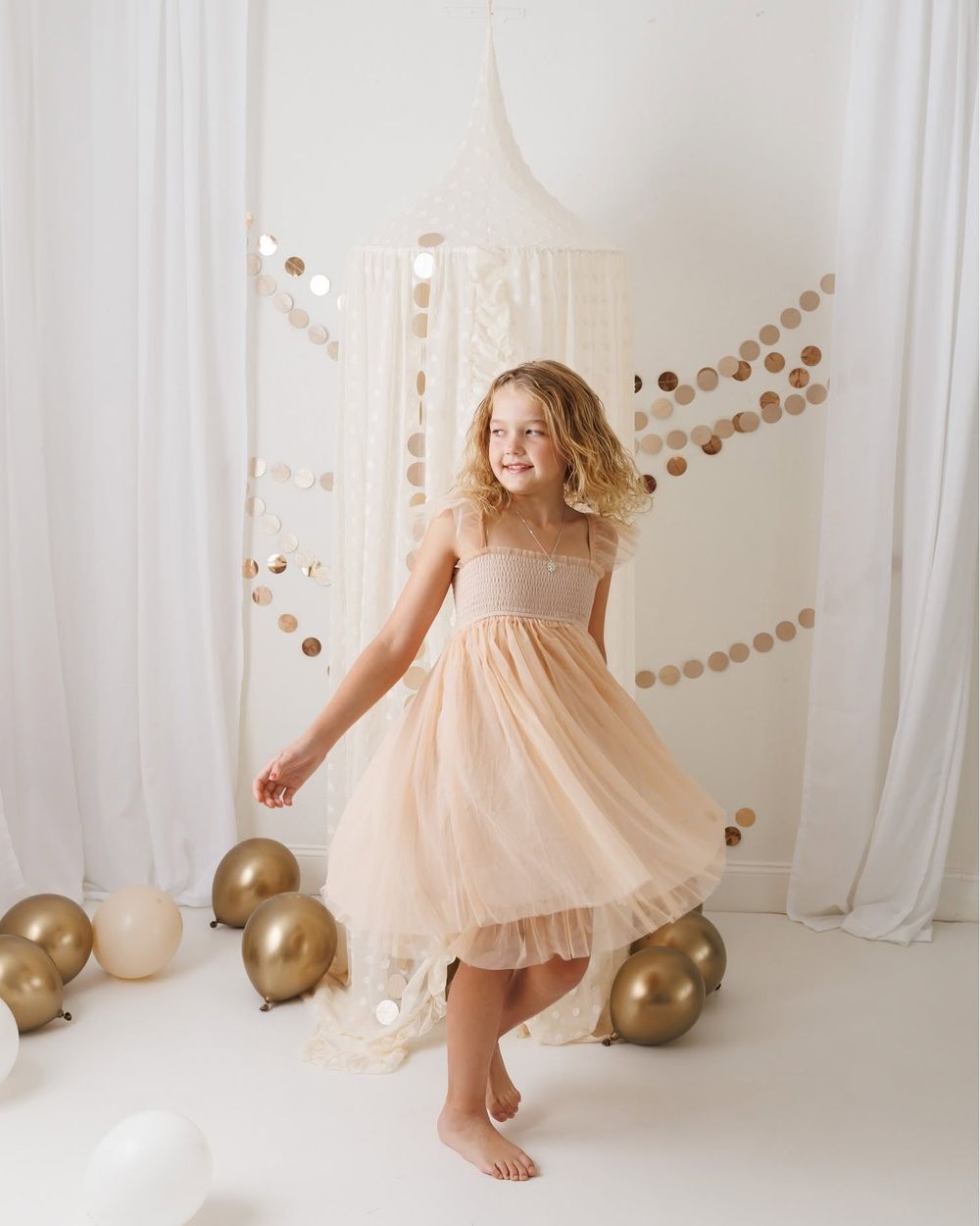 A young girl twirls in a peach tulle dress surrounded by gold balloons and circle garlands in a bright white studio.