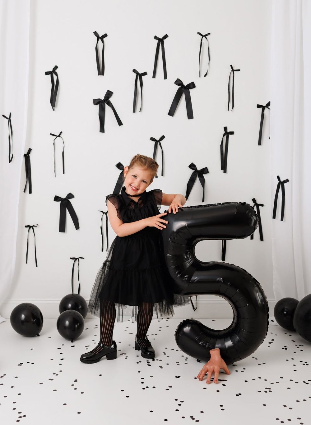 Girl in black dress poses with large number 5 balloon at birthday party with black bows and balloons decor.