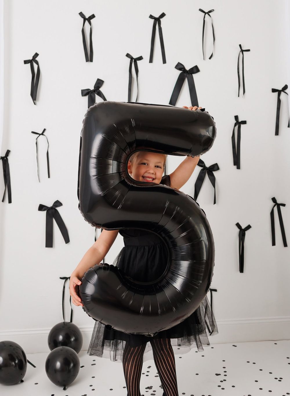 Smiling child holding large black number 5 balloon at birthday party with black bows on white wall backdrop.