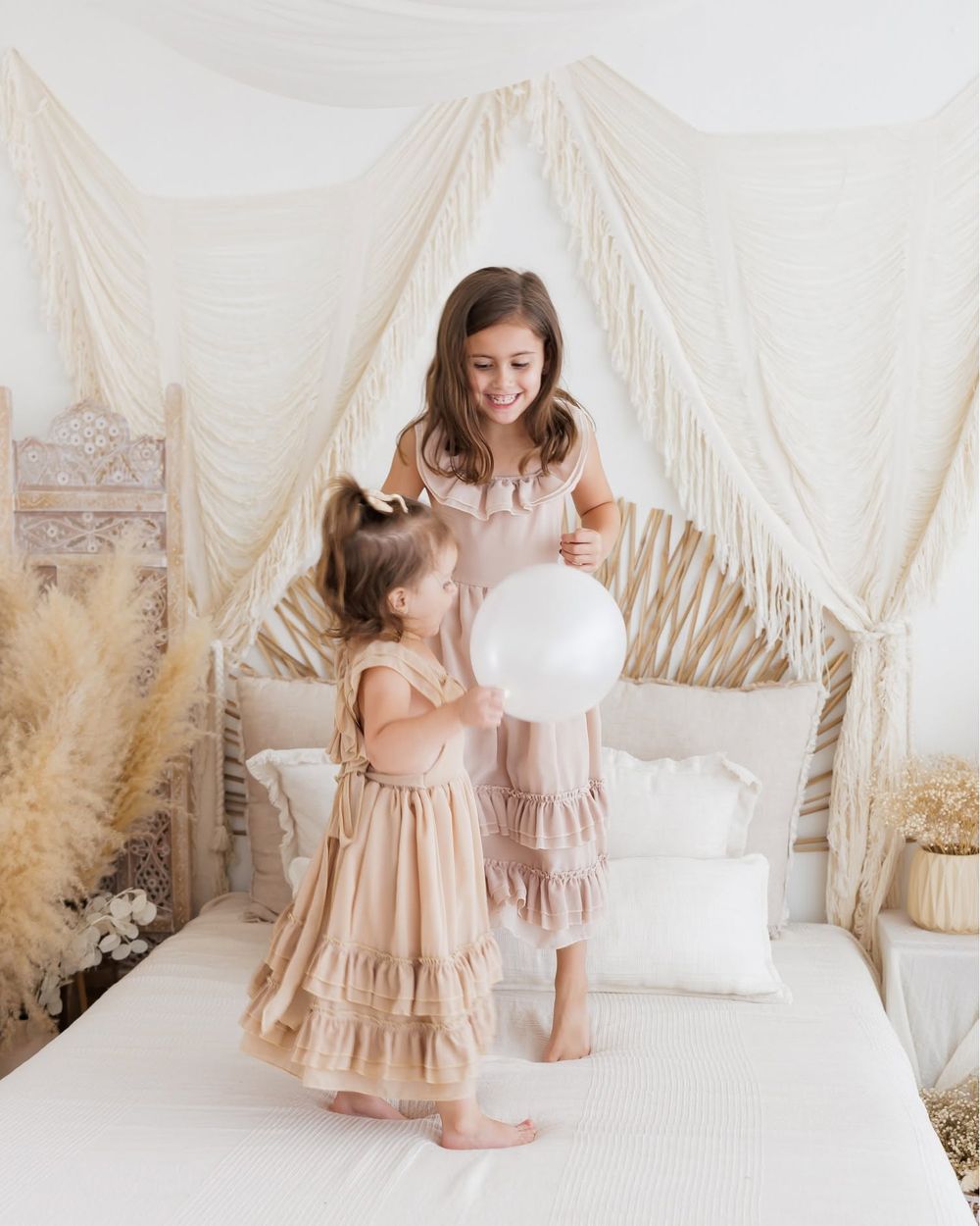 Two young girls in matching beige ruffle dresses play with a white balloon on a bed with boho macrame decor.