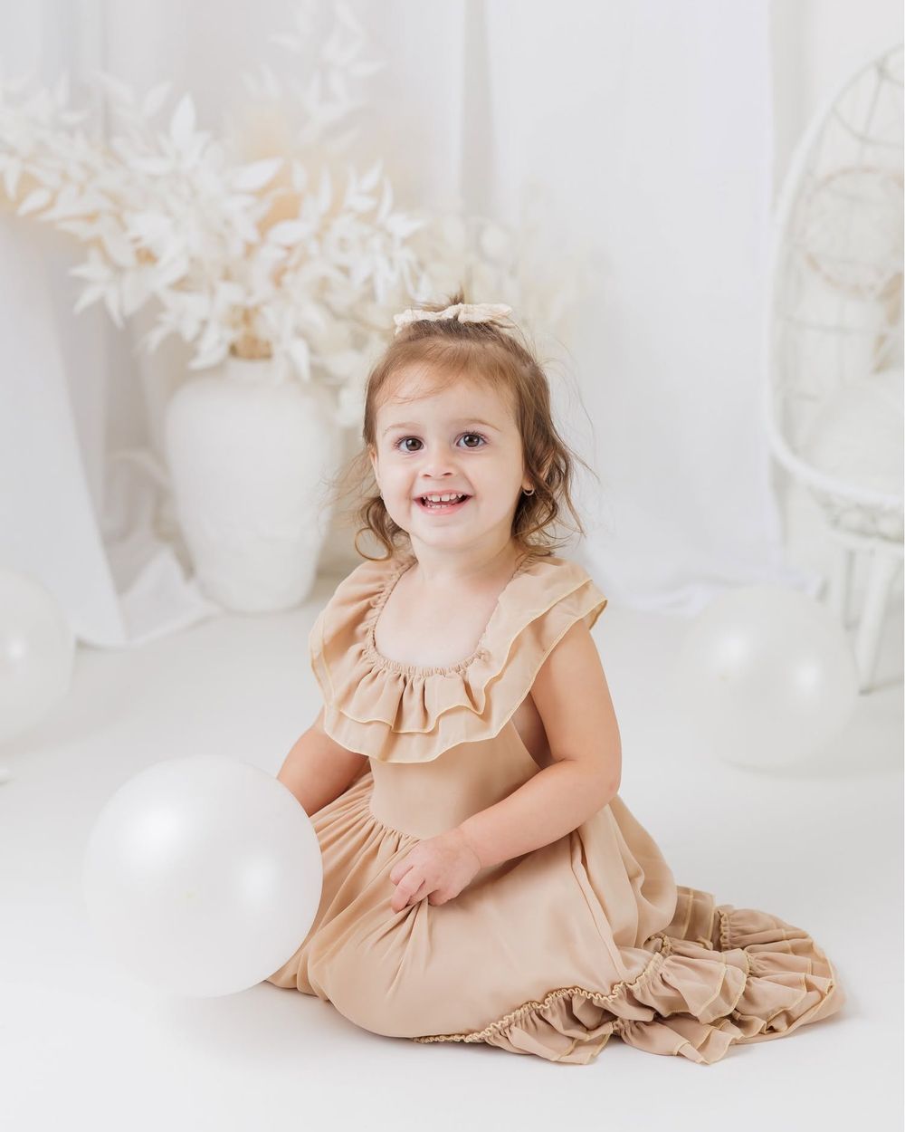 A smiling toddler girl in a tan ruffle dress sits on a white floor with a balloon in a bright studio setting.