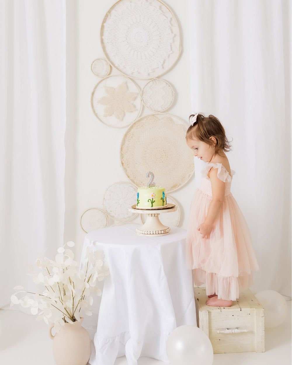 Toddler girl in pink tutu dress admires birthday cake on white table with boho dreamcatcher wall decor backdrop.