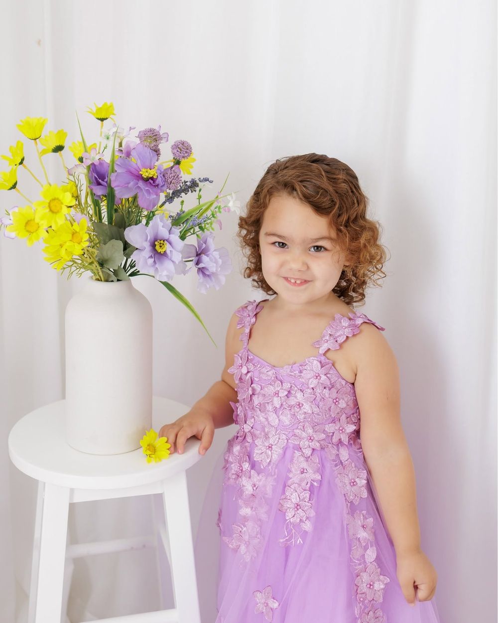 A young girl in a lavender floral dress smiles beside a white vase of purple and yellow flowers.