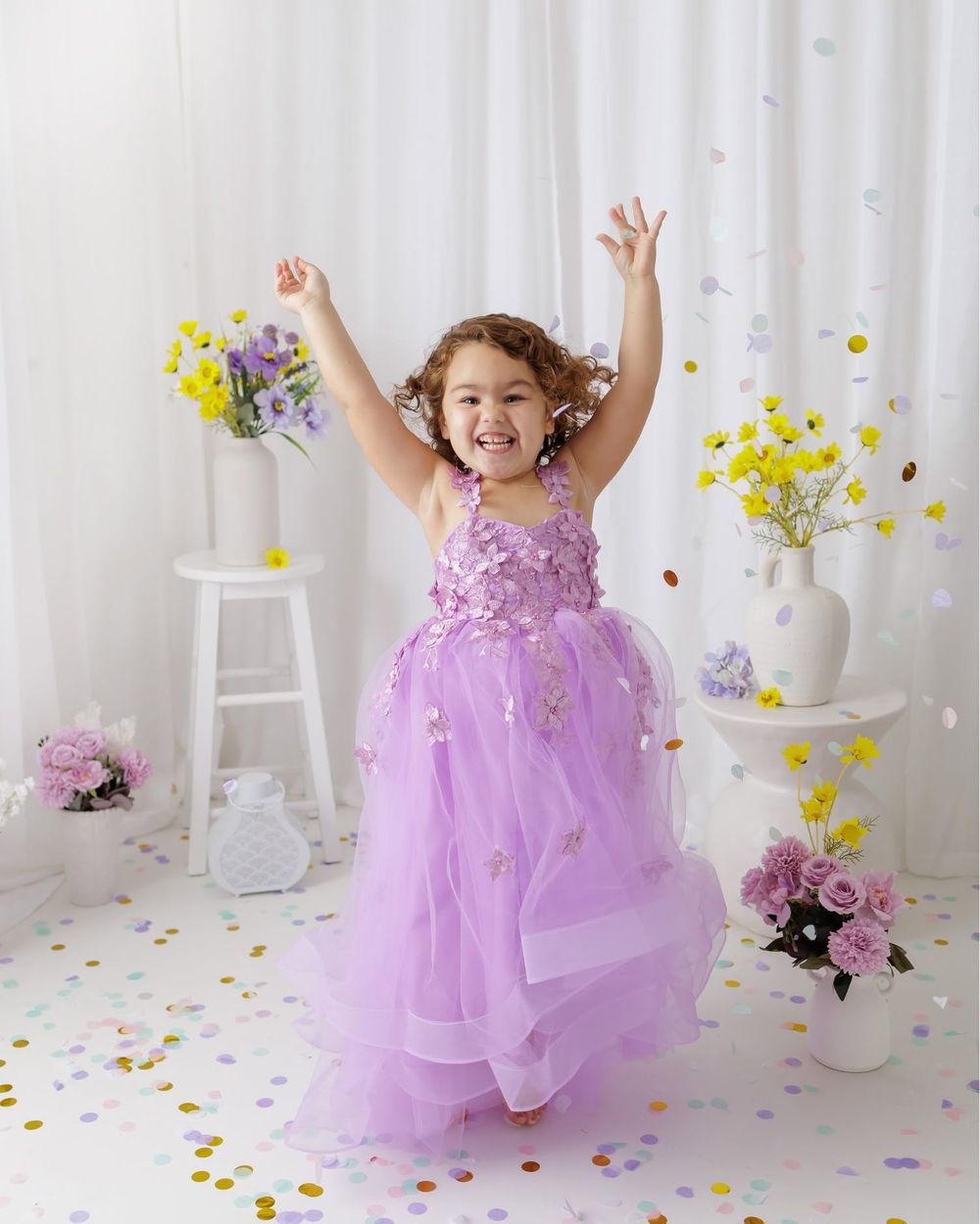 Joyful young girl in lavender tulle floral gown with arms raised, surrounded by confetti and flowers.
