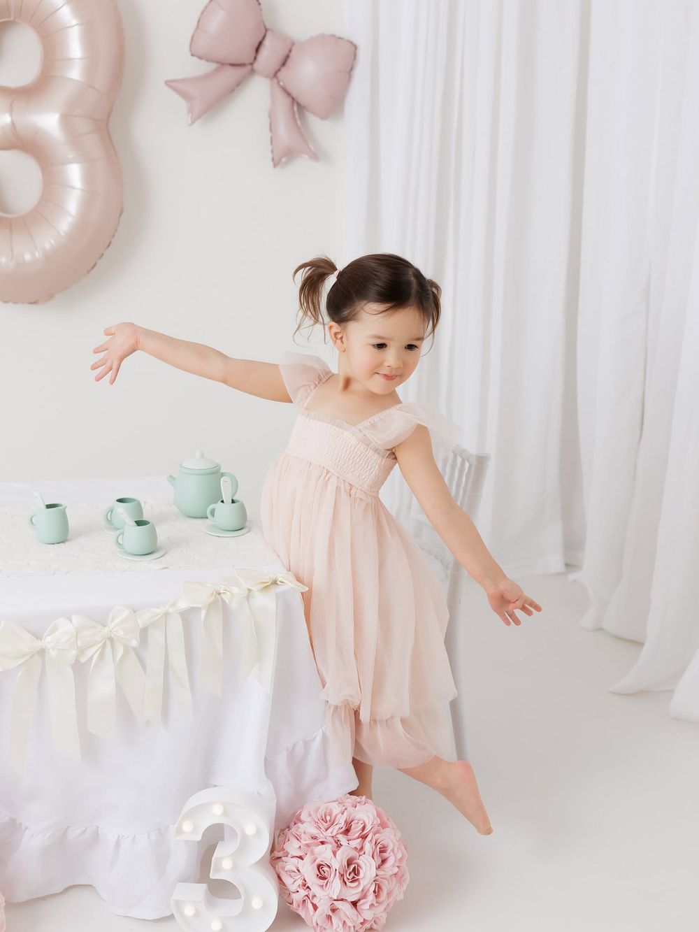 A young girl in a pink tulle dress poses playfully at a pastel tea party setup with a number 3 balloon for her birthday.