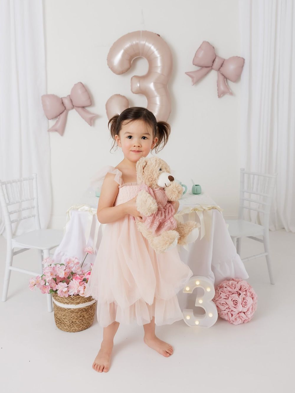A little girl in a pink tulle dress holds a teddy bear at her third birthday party with balloon decorations.