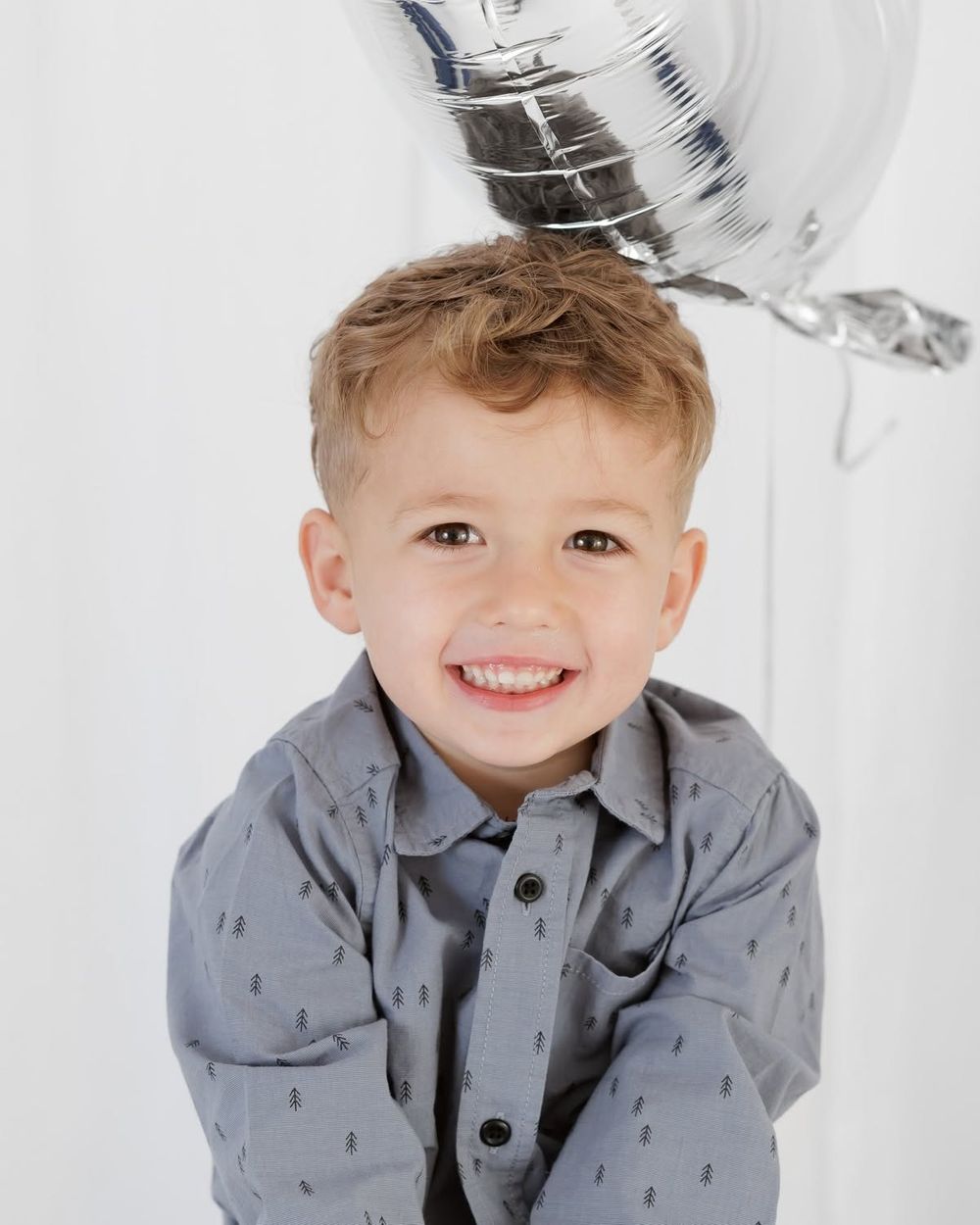 Smiling young boy in gray patterned button-up shirt posing with silver star balloon on white background.