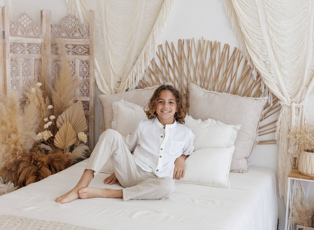 A smiling child sits cross-legged on a boho-style white bed with a palm leaf headboard and cream canopy.