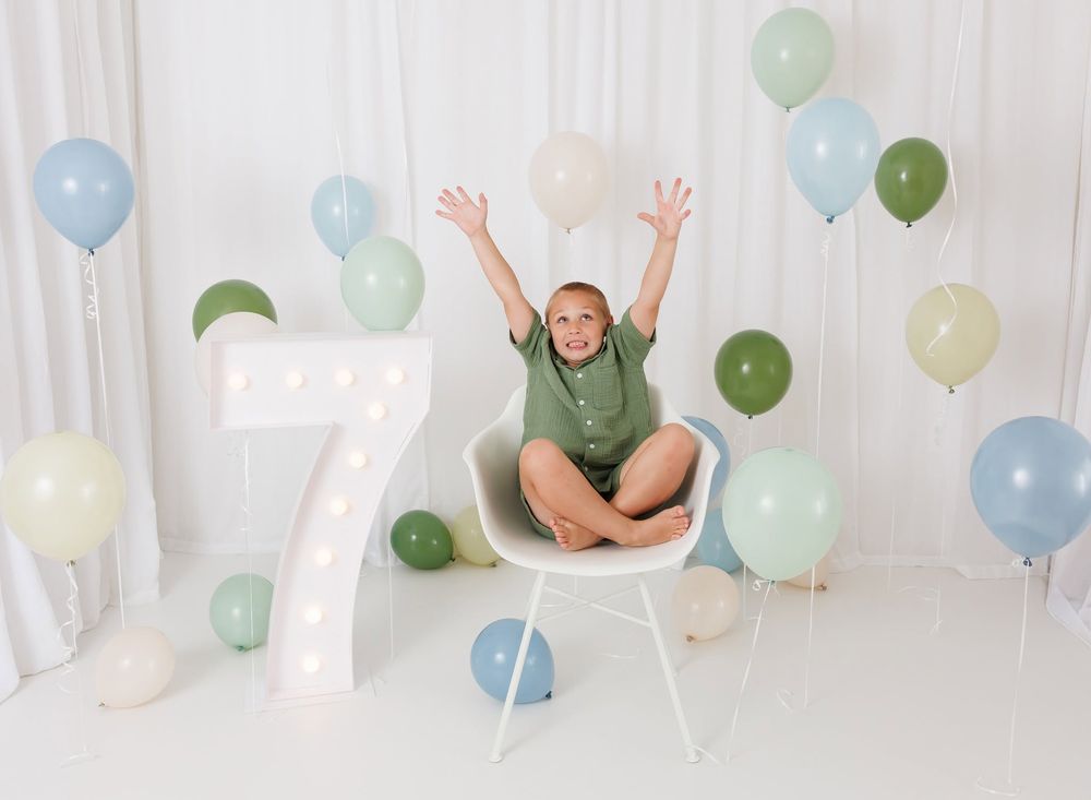 A joyful child celebrates turning 7, sitting on a chair with arms raised among green and blue balloons.