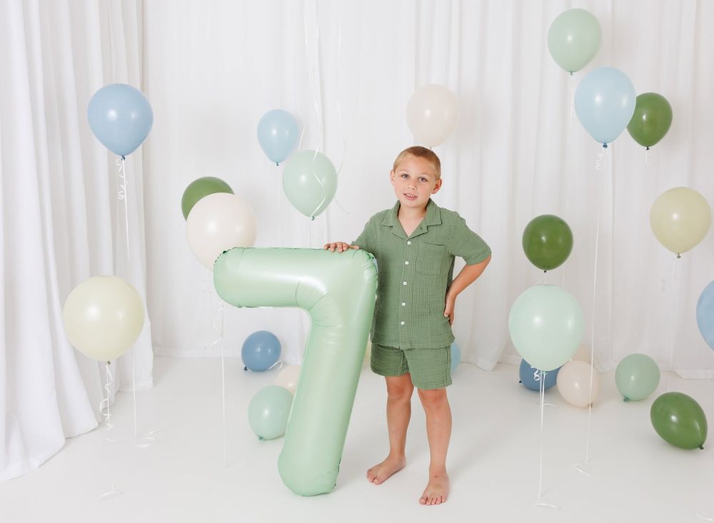 A young boy holds a large green number 7 balloon surrounded by pastel balloons for a 7th birthday photo.