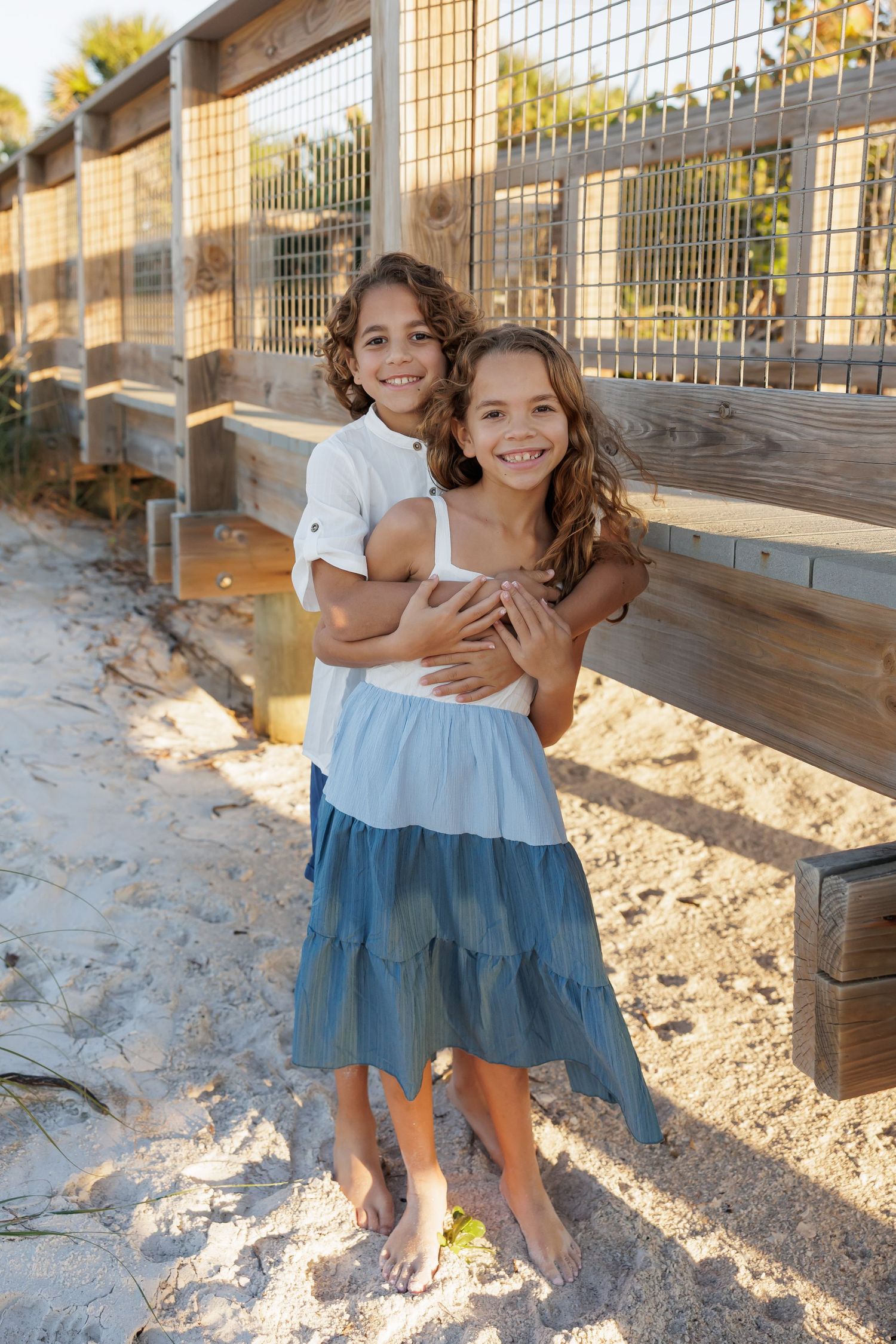 Two smiling children hugging on a sandy beach path beside a wooden fence on a sunny day.