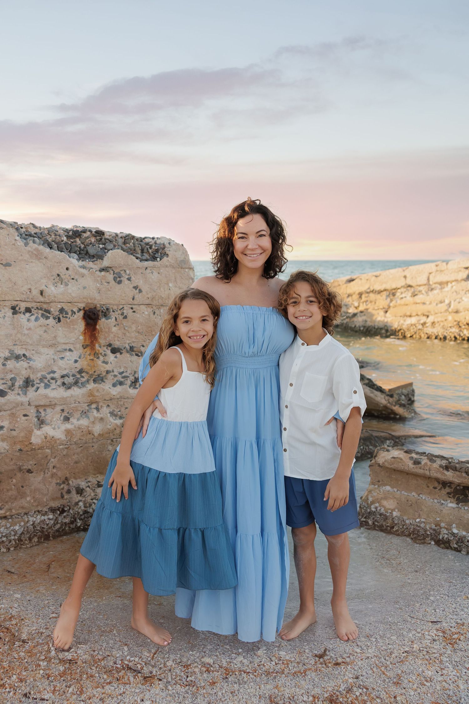 A woman in a blue dress poses with two children by rocky seaside ruins at sunset.