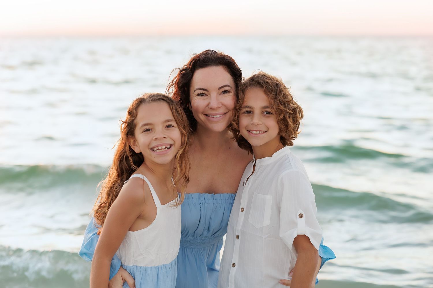 A smiling mother and two children pose together on a sandy beach with ocean waves in the background.