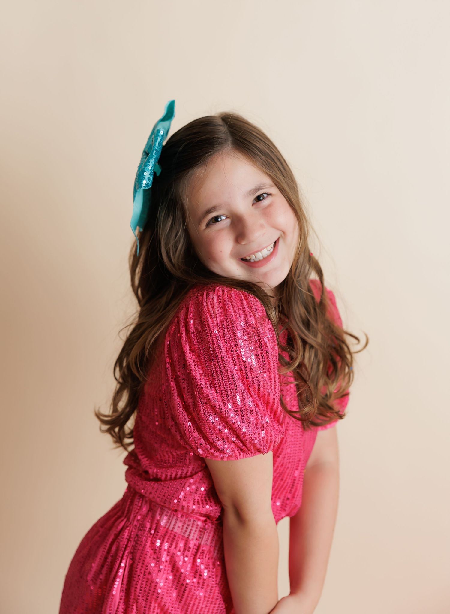 Smiling young girl in pink sequin dress with teal bow hair accessory posing against beige background.