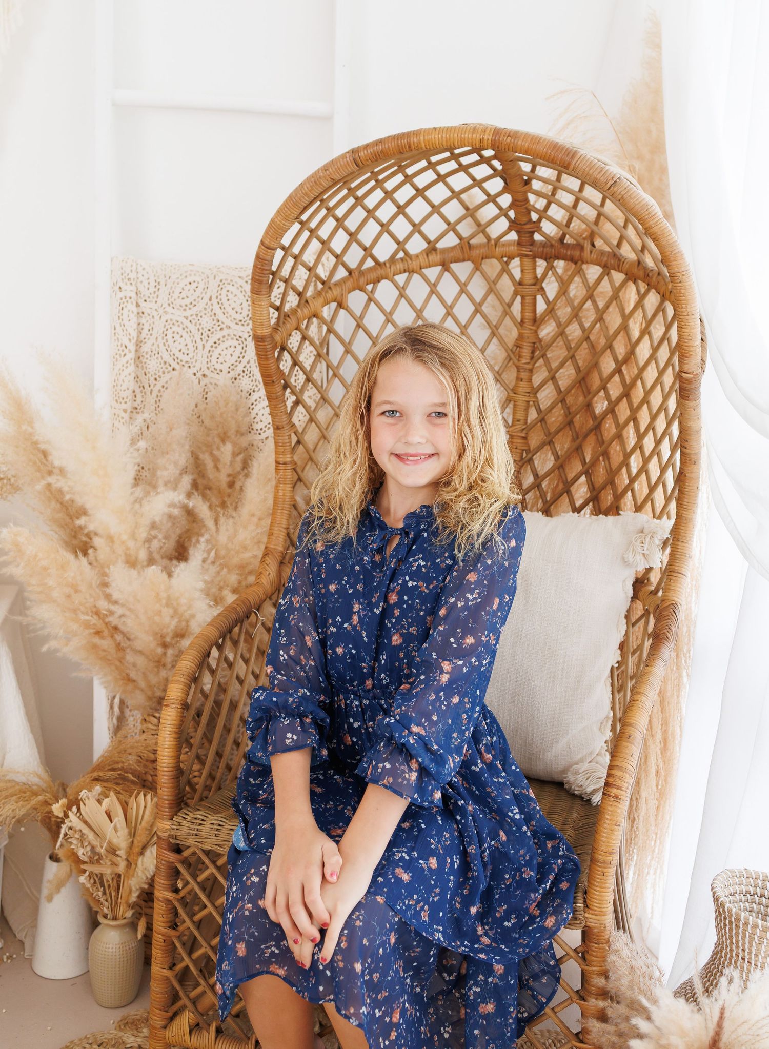 Young girl in blue floral dress sitting in a wicker peacock chair with pampas grass decor.