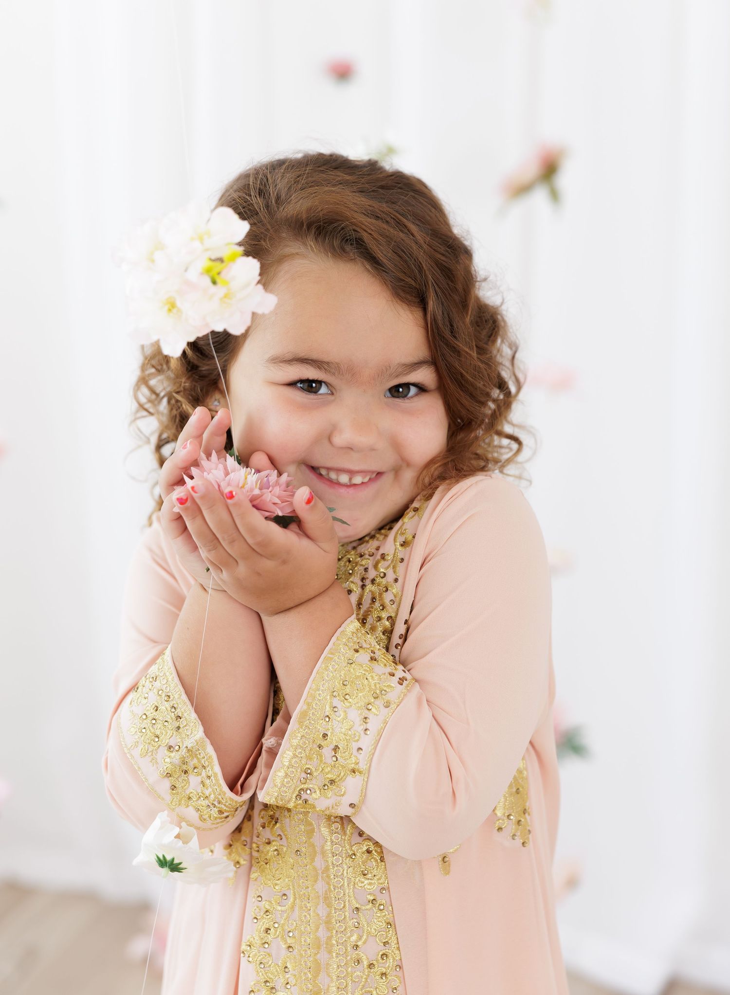 A smiling young girl with curly hair and a flower accessory holds pink blooms in her hands, wearing a blush and gold dress.