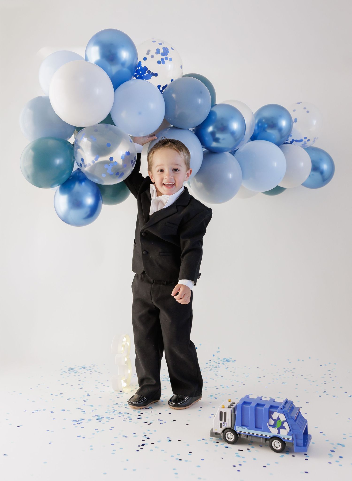 Young boy in black suit holding blue balloon garland smiles beside a toy recycling truck with confetti on white background.