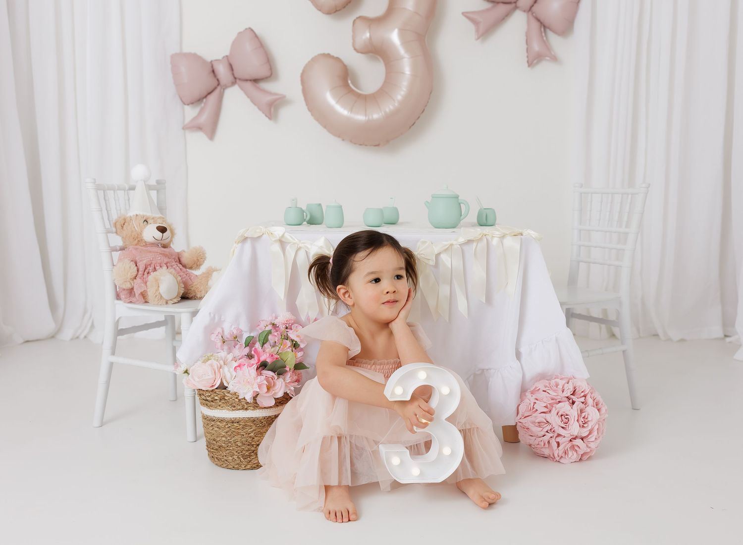 Toddler girl in pink tutu sits with number 3 balloon and decorations at a pastel birthday party setup.