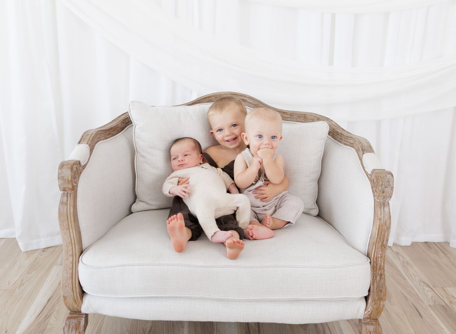 Three babies sitting together on a vintage cream sofa against a white backdrop in a professional photo studio setting.