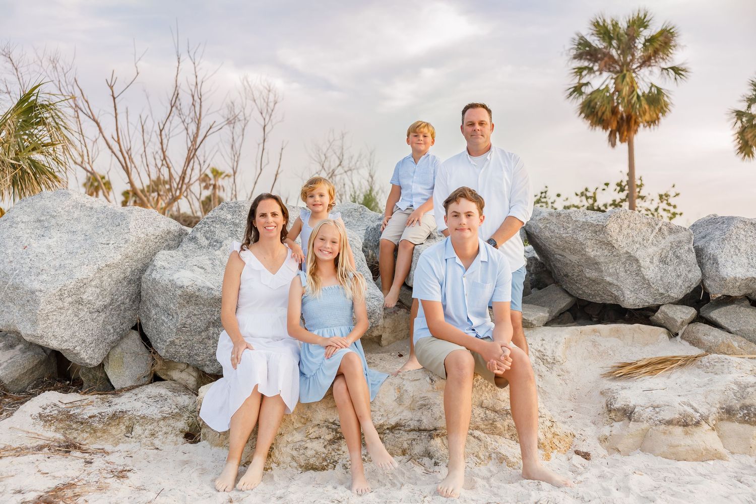 Family of five posing on rocky beach with palm trees, dressed in coordinating white and blue outfits for a portrait.