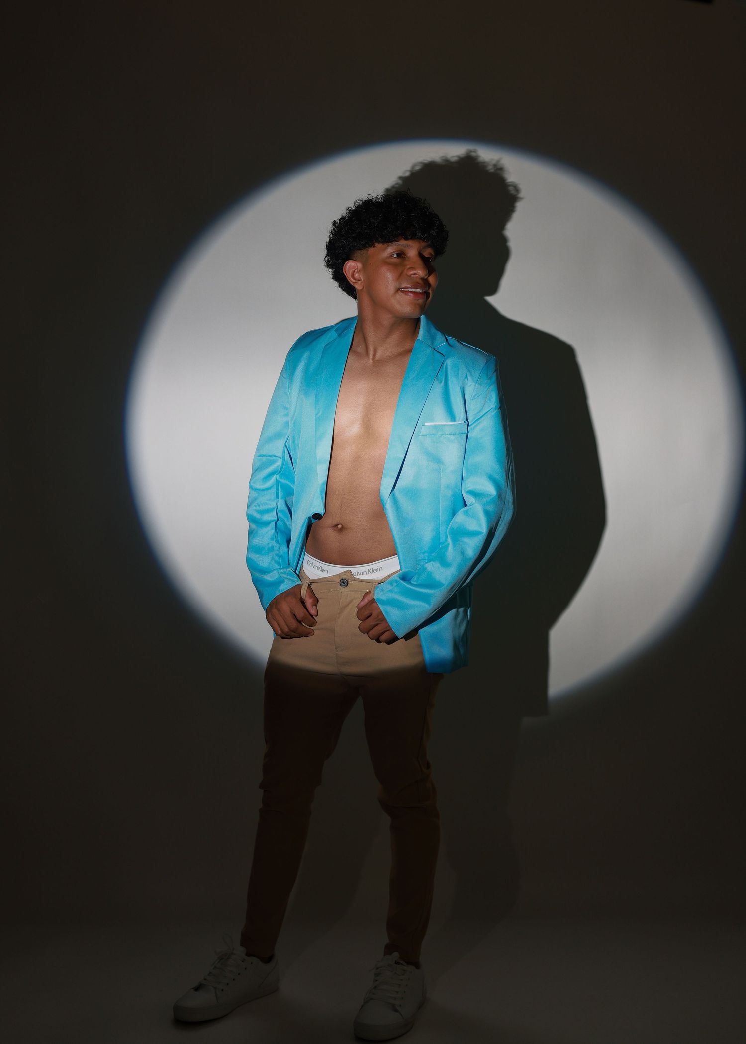 Young man posing in cyan blazer, open chest, dark jeans, white sneakers under dramatic spotlight on gray background.