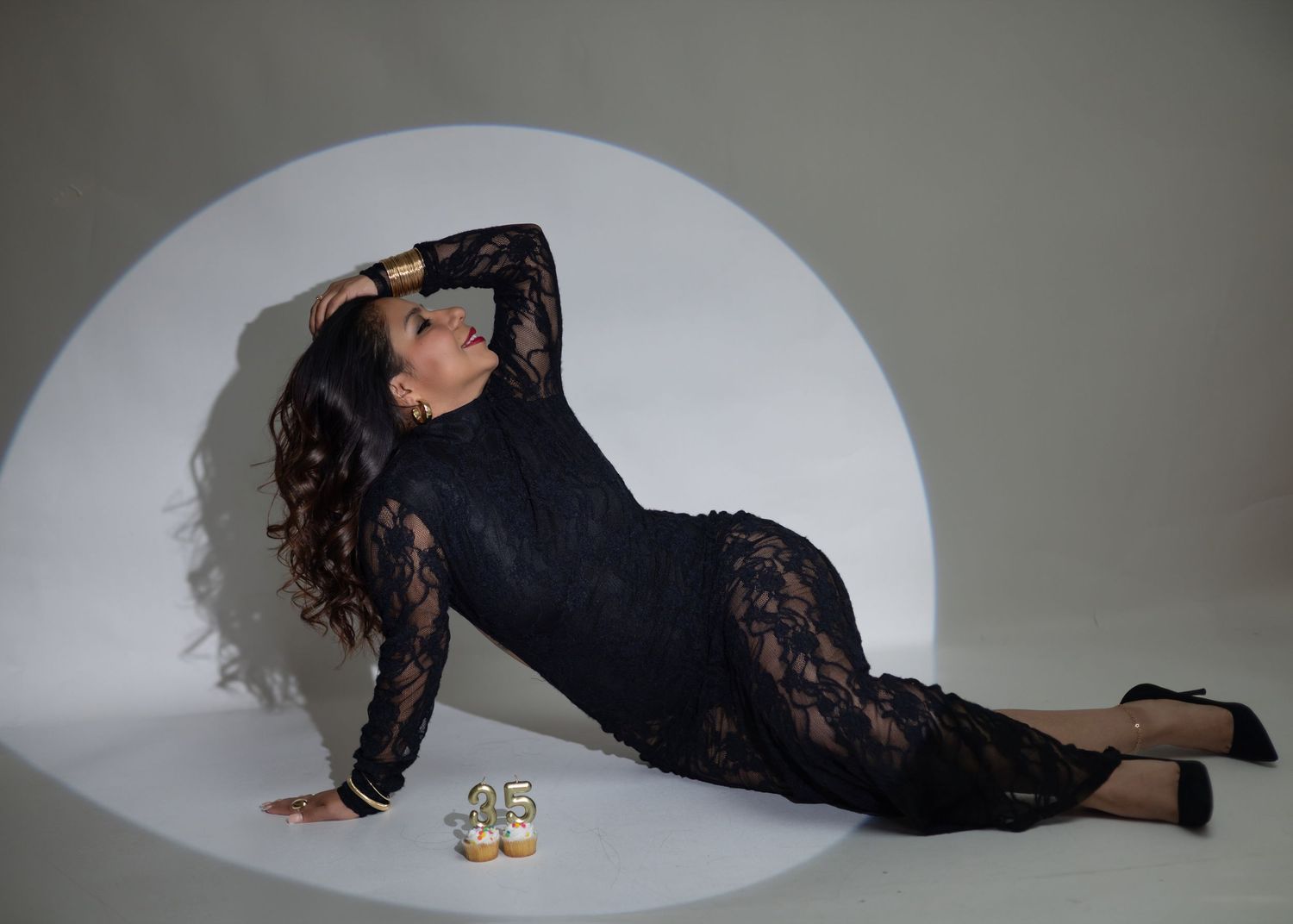 Woman in black lace dress posing on floor with gold cuffs and heels, celebrating 35th birthday.