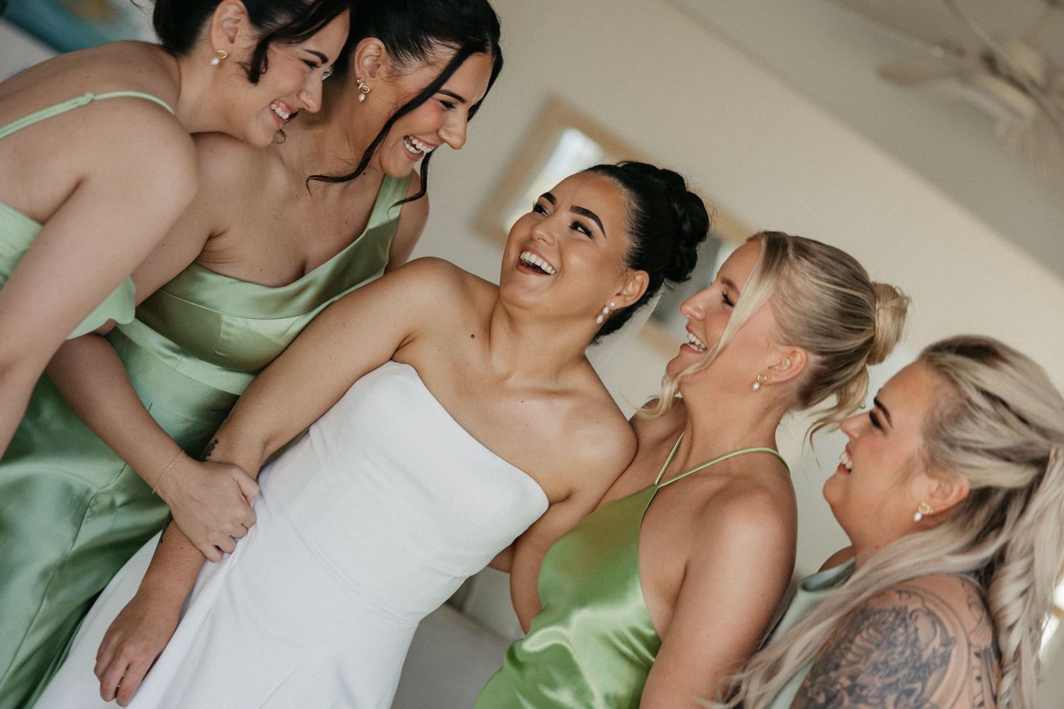Bride in white strapless gown laughing with bridesmaids in sage green satin dresses during wedding preparations.