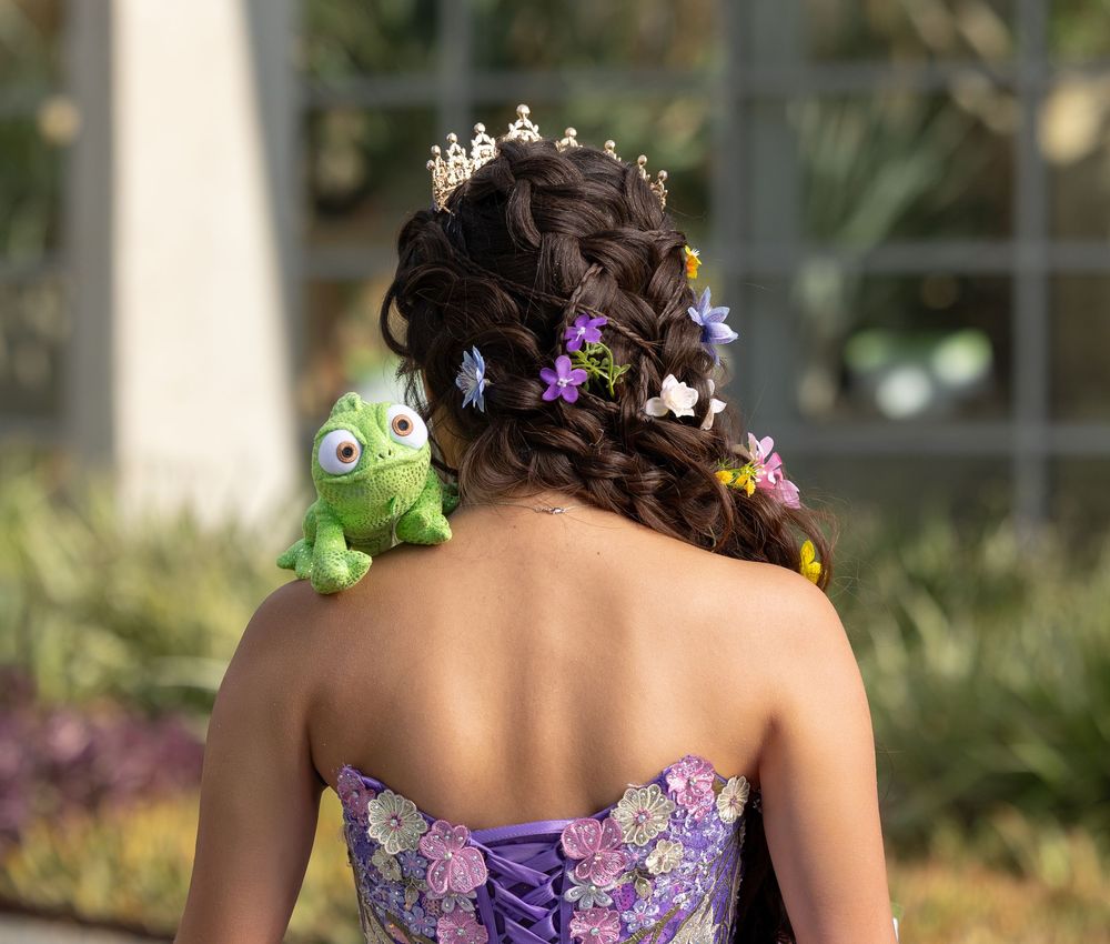 Young woman in purple floral strapless gown with braided updo, tiara, flowers, and Pascal chameleon toy on shoulder.