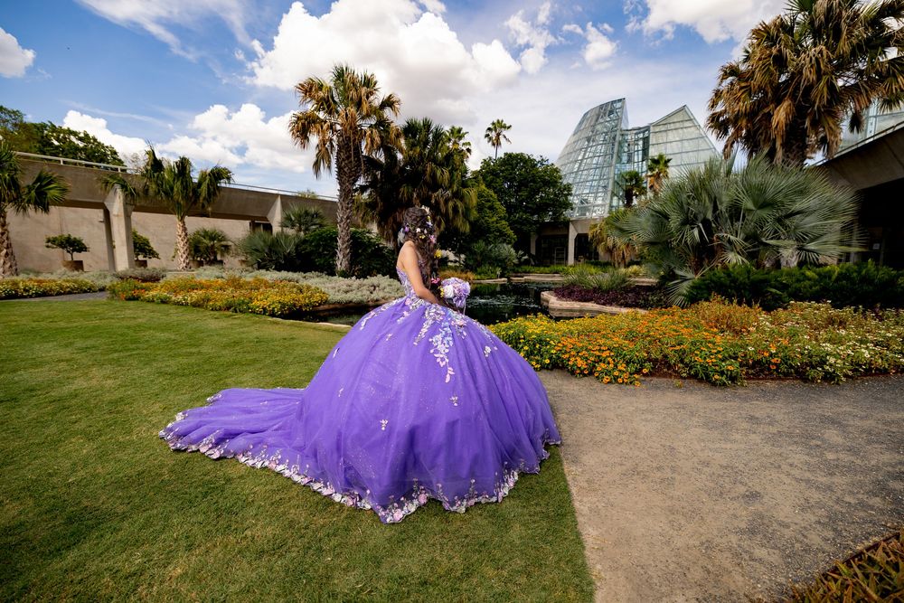 Girl in voluminous purple quinceañera ball gown poses in lush garden with palm trees and glass building backdrop.