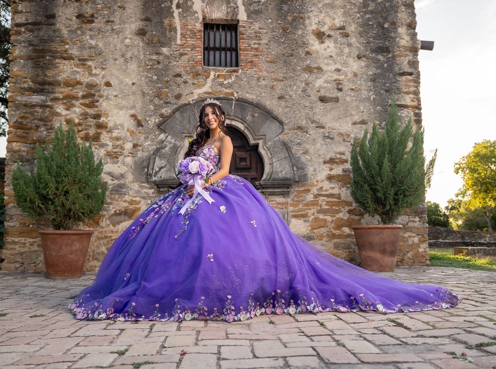 Quinceañera in purple ball gown with floral details poses before historic stone building.