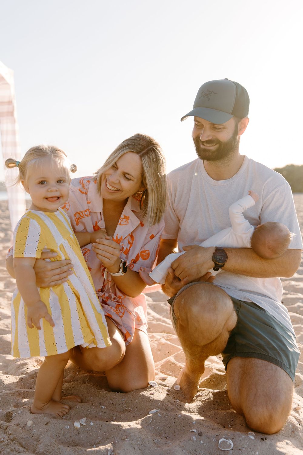 Happy family with toddler girl and newborn baby posing together on sandy beach in warm golden sunlight.