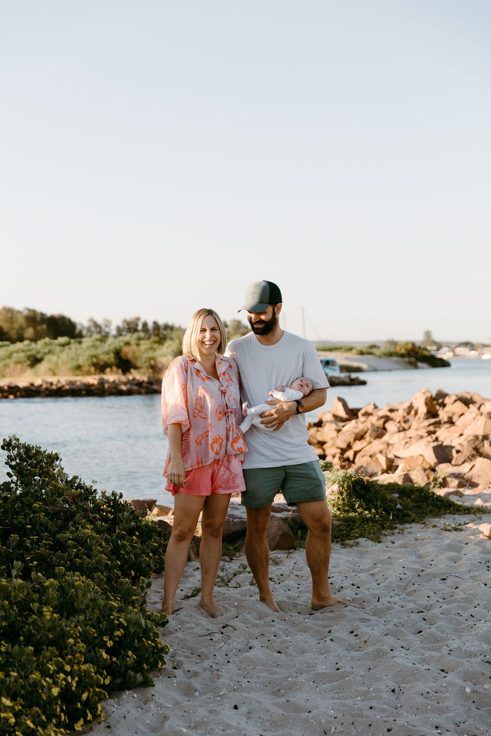 A couple stands barefoot on a sandy beach holding a newborn baby, with rocky shoreline and water in the background.