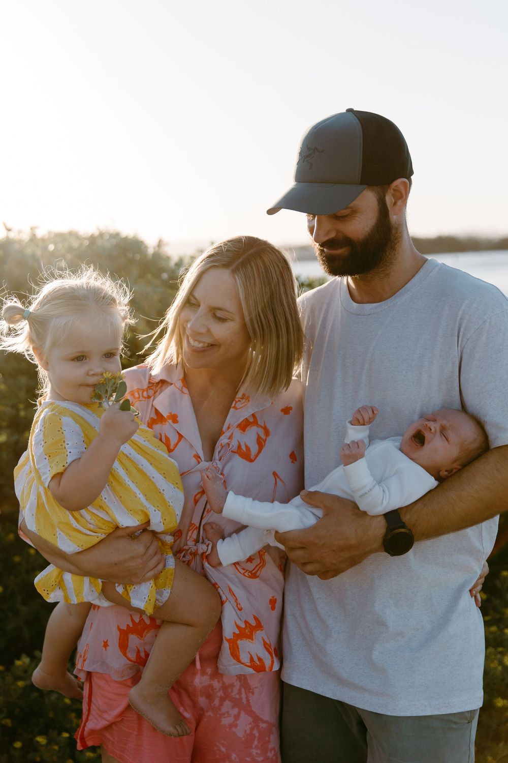 Young family with toddler and newborn baby posing outdoors at golden hour near waterfront.