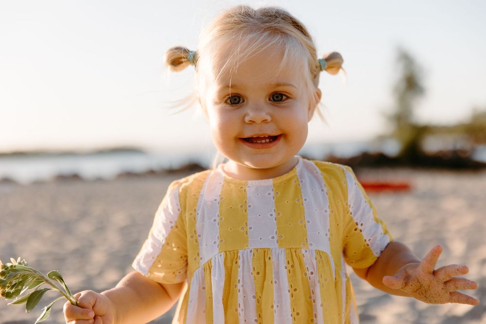 Smiling toddler girl in yellow striped dress playing on sandy beach at sunset.