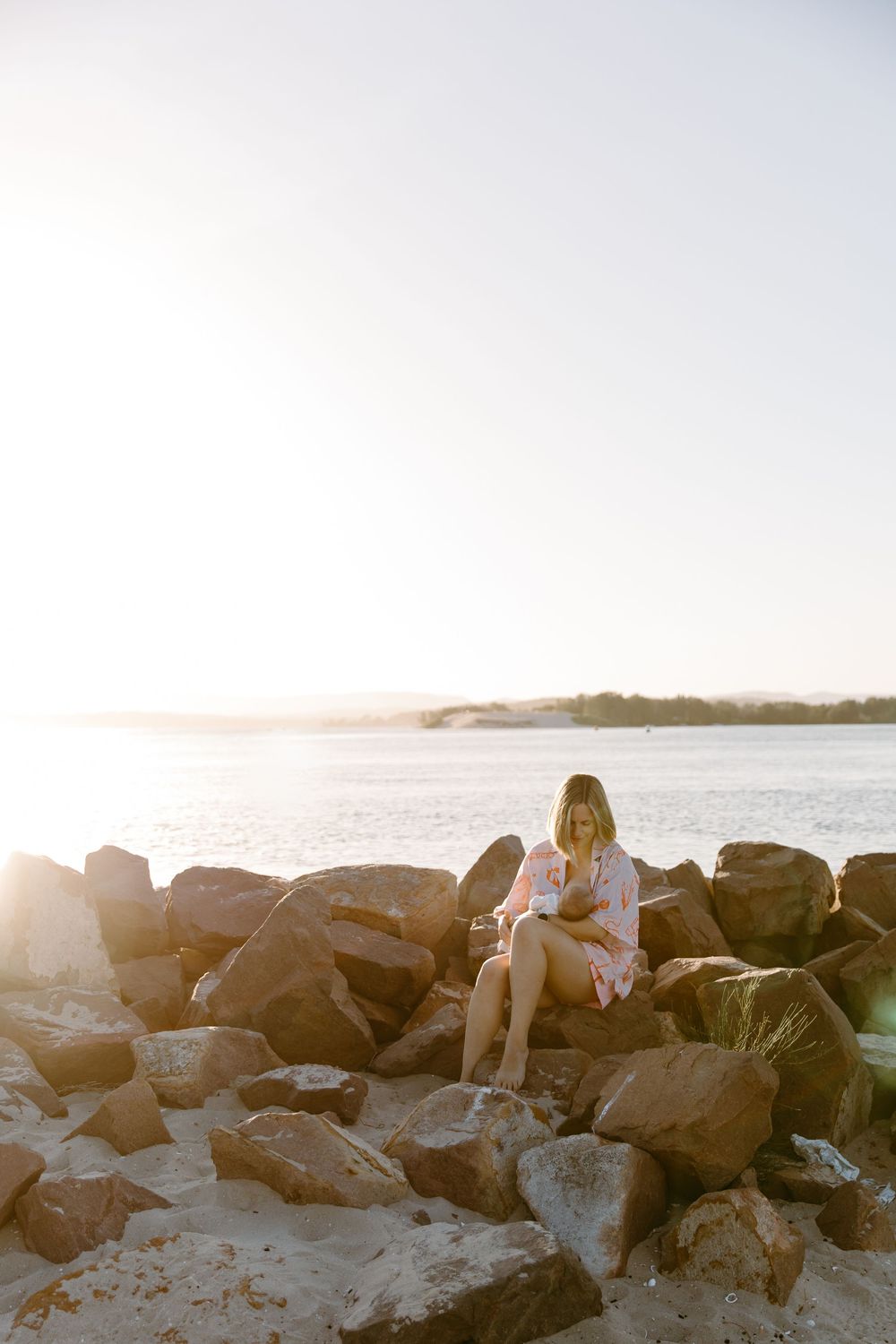 Woman sitting on rocky shoreline at golden hour sunset with calm ocean waters in the background.