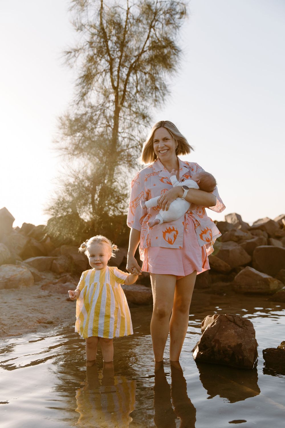 A smiling mother in a pink outfit holds a newborn while walking in shallow water with her toddler in a yellow dress.