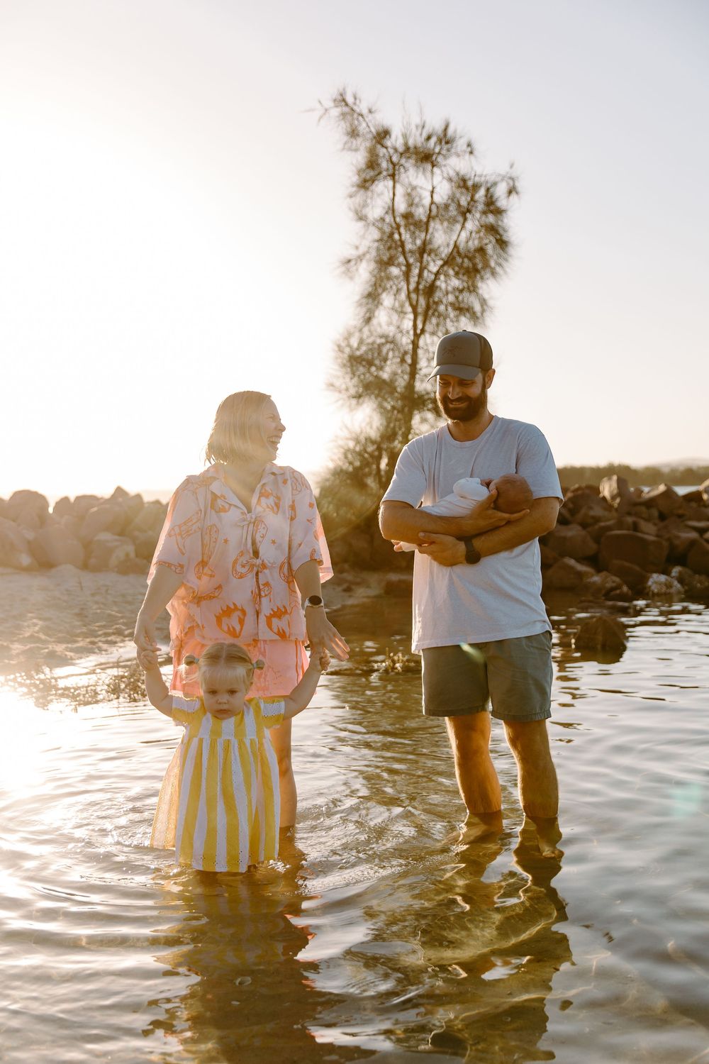 Family of three wading in shallow water at sunset, parents holding children near rocky shore.