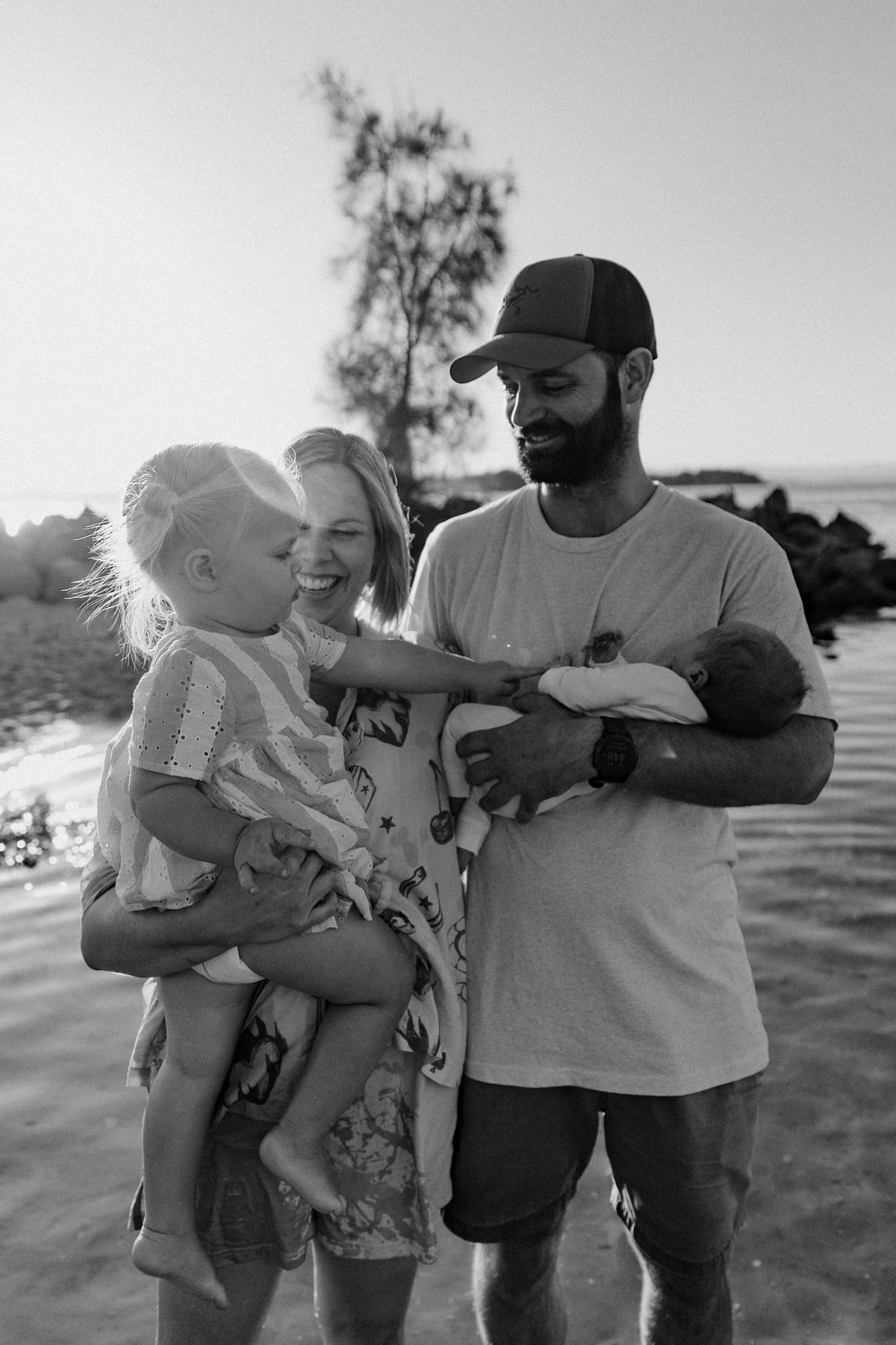 Black and white photo of a happy family at the beach, parents holding two young children, smiling together.