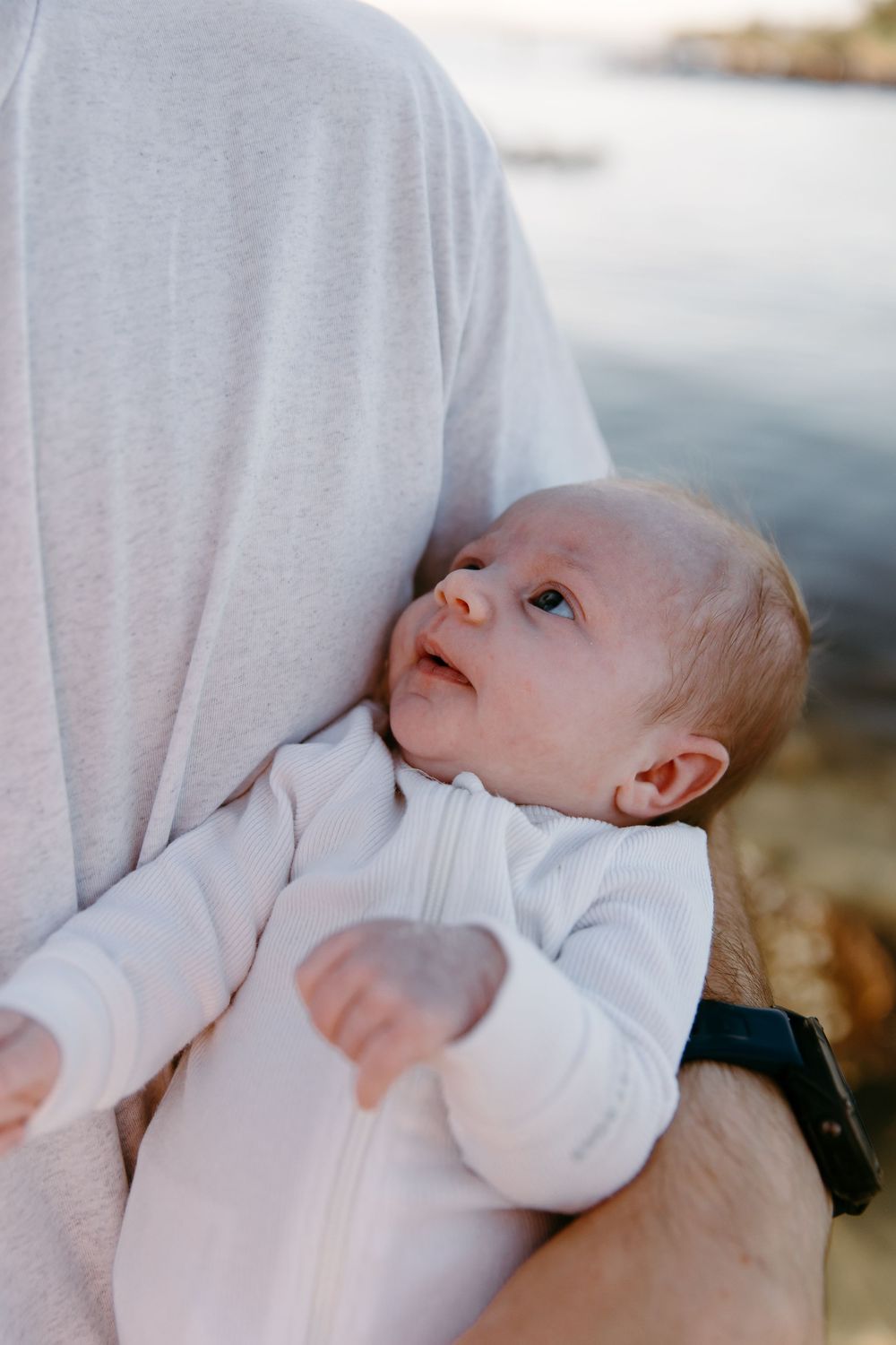 A newborn baby in a white zip-up onesie being cradled in an adult's arms near a serene waterfront.