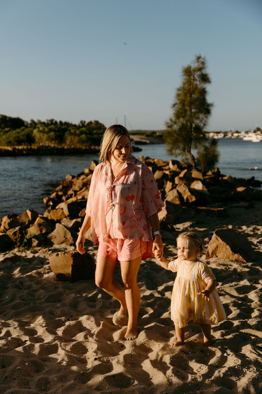 A mother in pink holds her toddler's hand on a sandy beach near rocky shores at golden hour.