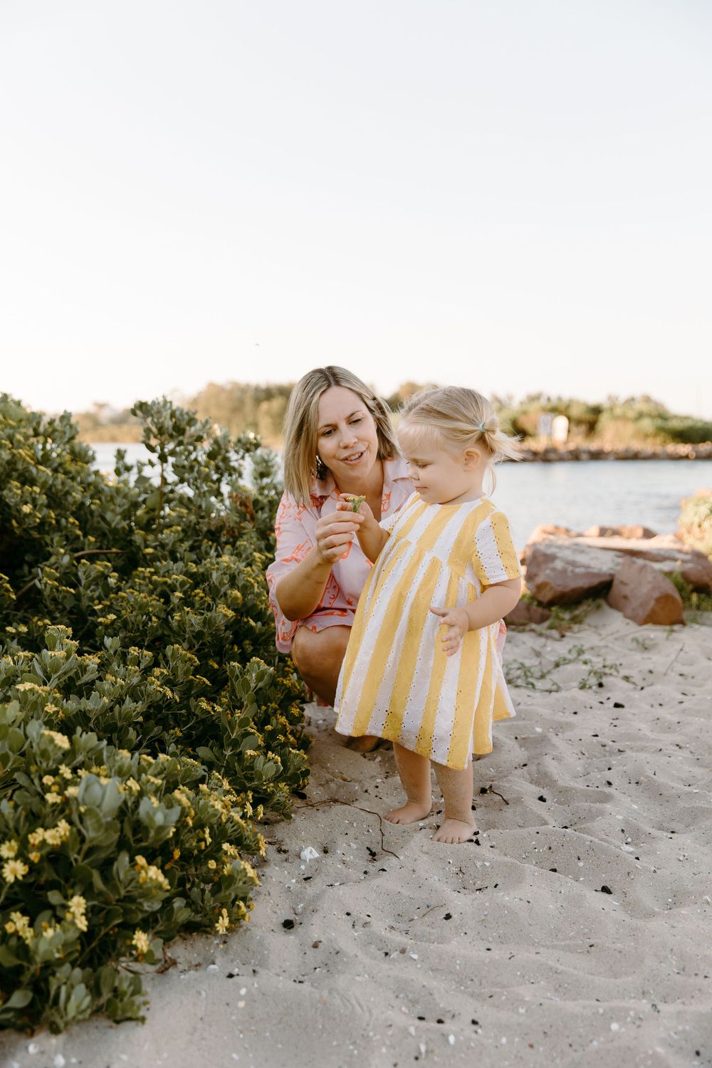 A mother crouches beside her young daughter in a yellow striped dress on a sandy beach, exploring coastal plants together.