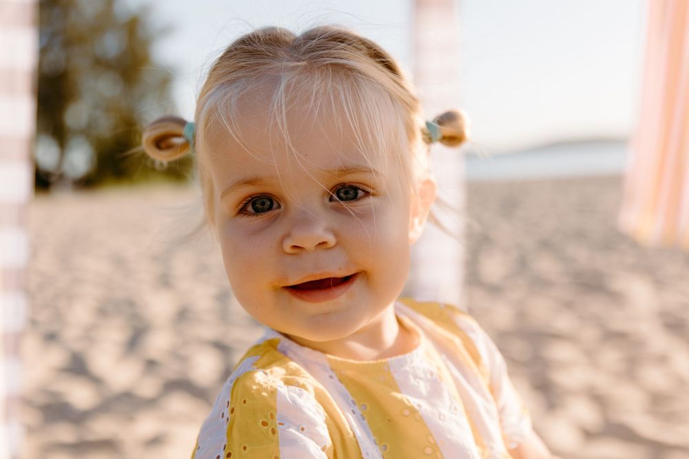 Adorable smiling toddler girl with pigtails at a sunny beach, wearing a yellow outfit.
