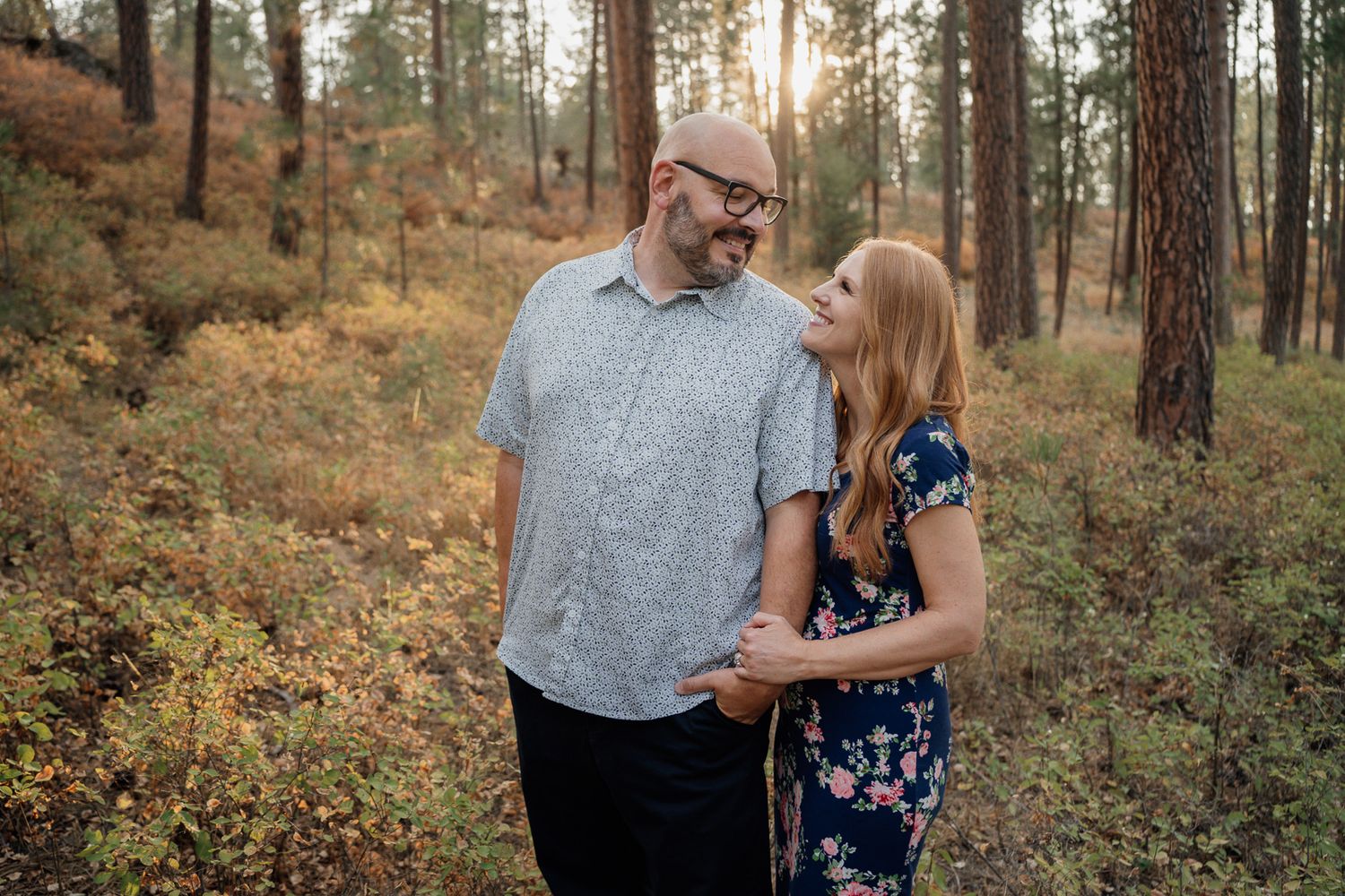 Family of four smiling together in a sunlit pine forest during golden hour, wearing casual and floral outfits.