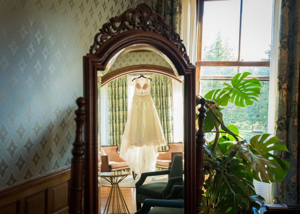 Elegant wedding dress hanging in ornate wooden mirror reflection near bright window in decorated room.