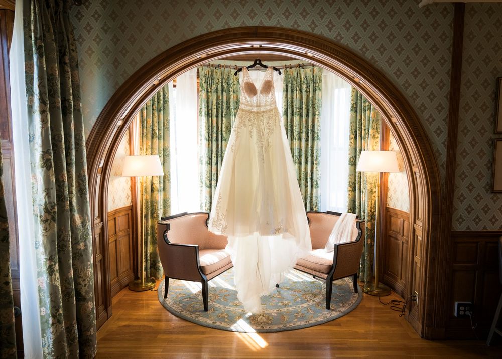 Elegant wedding dress displayed under a wooden arch in a warmly lit bridal suite with green curtains.