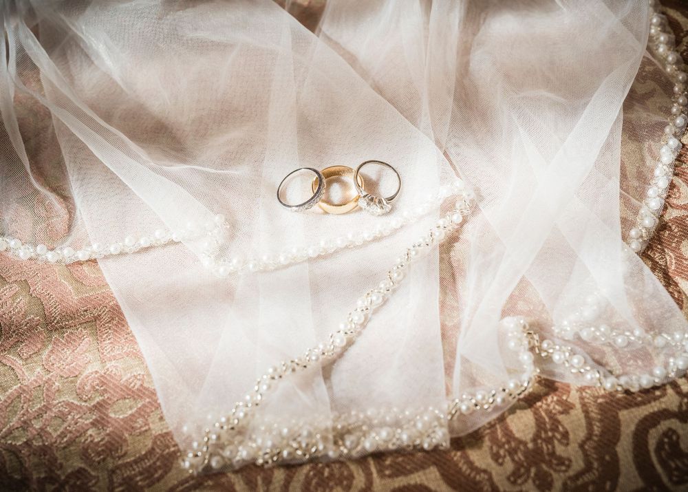 Wedding rings and pearl jewelry resting on a white lace bridal veil with decorative fabric underneath.