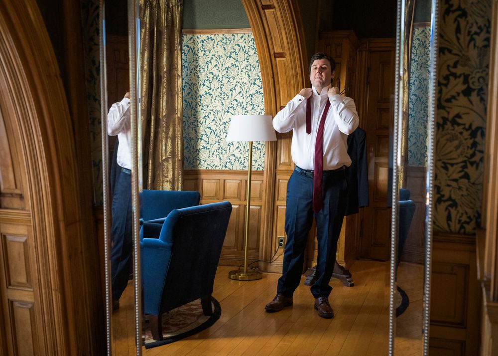 Man in white shirt and red tie adjusts collar before mirror in elegant wood-paneled room with blue chair.