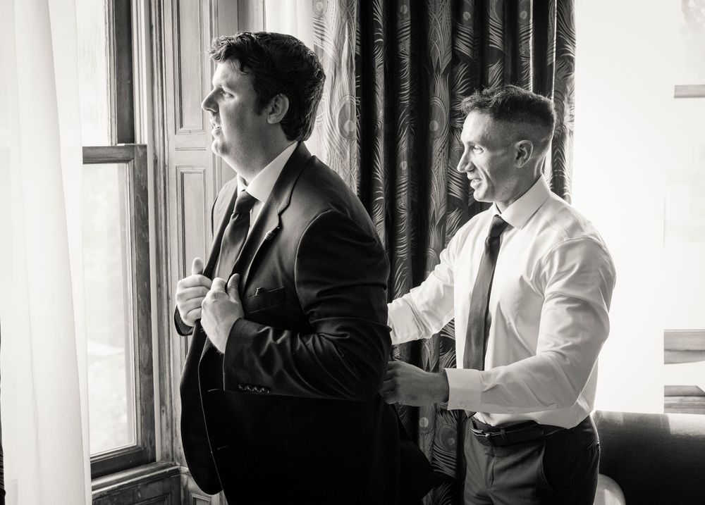 Two men in formal suits assist each other getting dressed in black and white wedding preparation photo.